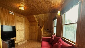 Interior de uma cabana de madeira com televisão, sofá vermelho, escadas de madeira e janelas amplas.