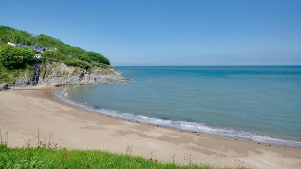 A beach with a sandy shore, a grassy foreground, and a cliff with buildings on top. - 3 Bed in Aberporth (oc-g27666) (Aberporth)