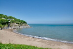 A beach with a sandy shore, a grassy foreground, and a cliff with buildings on top. - 3 Bed in Aberporth (oc-g27666) (Aberporth)