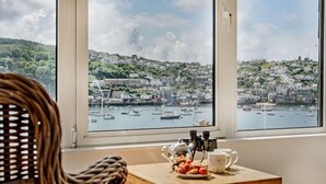 Une pièce avec vue sur un port, une chaise en osier, une table en bois avec une théière et une assiette de fraises.