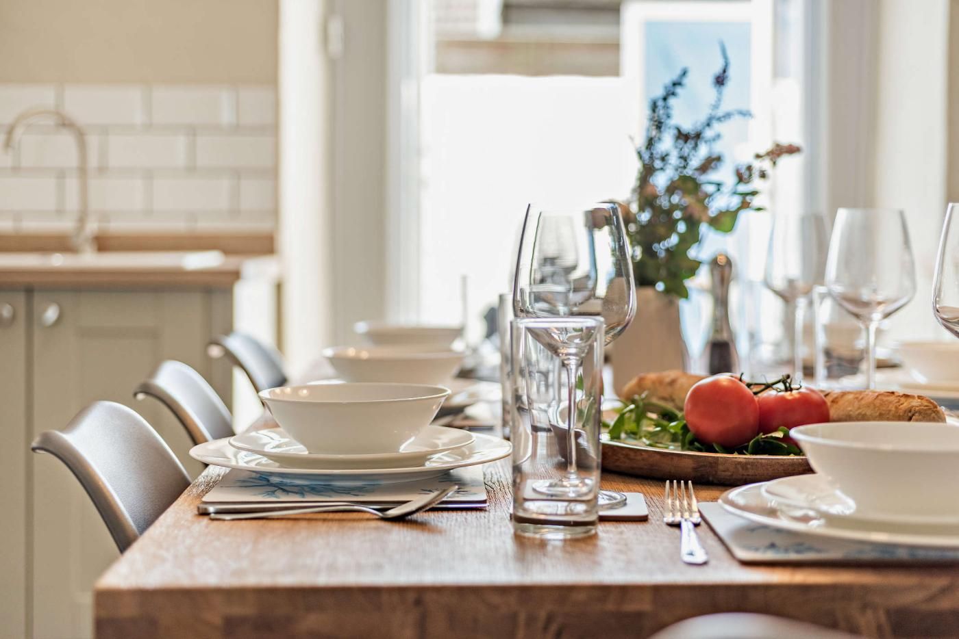 A dining table set with white plates, bowls, glasses, and cutlery. There is a bottle of water, a glass pitcher, and a wooden board with bread and tomatoes.