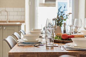 Une table à manger est dressée avec de l’assiette blanche, des bols, des verres et des couverts. On y trouve une bouteille d’eau, un pichet en verre et une planche en bois sur laquelle sont disposés du pain et des tomates.