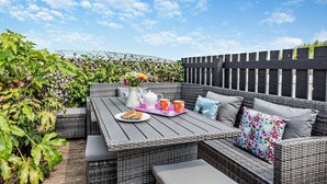 A wicker outdoor seating area with a table, cushions, and a plate of food. - 4 Bed in Westward Ho (oc-h27657) (Westward Ho!)