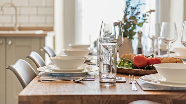 A dining table set with white plates, bowls, glasses, and cutlery. There is a bottle of water, a glass pitcher, and a wooden board with bread and tomatoes. - 4 Bed in Westward Ho (oc-h27657) (Westward Ho!)