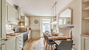 A modern kitchen with wooden cabinets, a central island, and a dining area with a long table and chairs. - 4 Bed in Westward Ho (oc-h27657) (Westward Ho!)