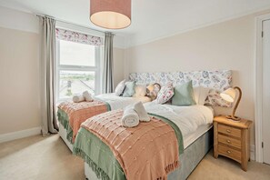A bedroom with two beds, a window with curtains, a bedside lamp, and a view of the outside. - 4 Bed in Westward Ho (oc-h27657) (Westward Ho!)