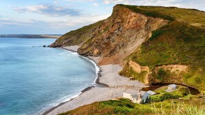 A coastal landscape with a beach, cliffs, and a small building with a chimney. - 1 Bed in Welcombe (oc-h27741) (Welcombe)