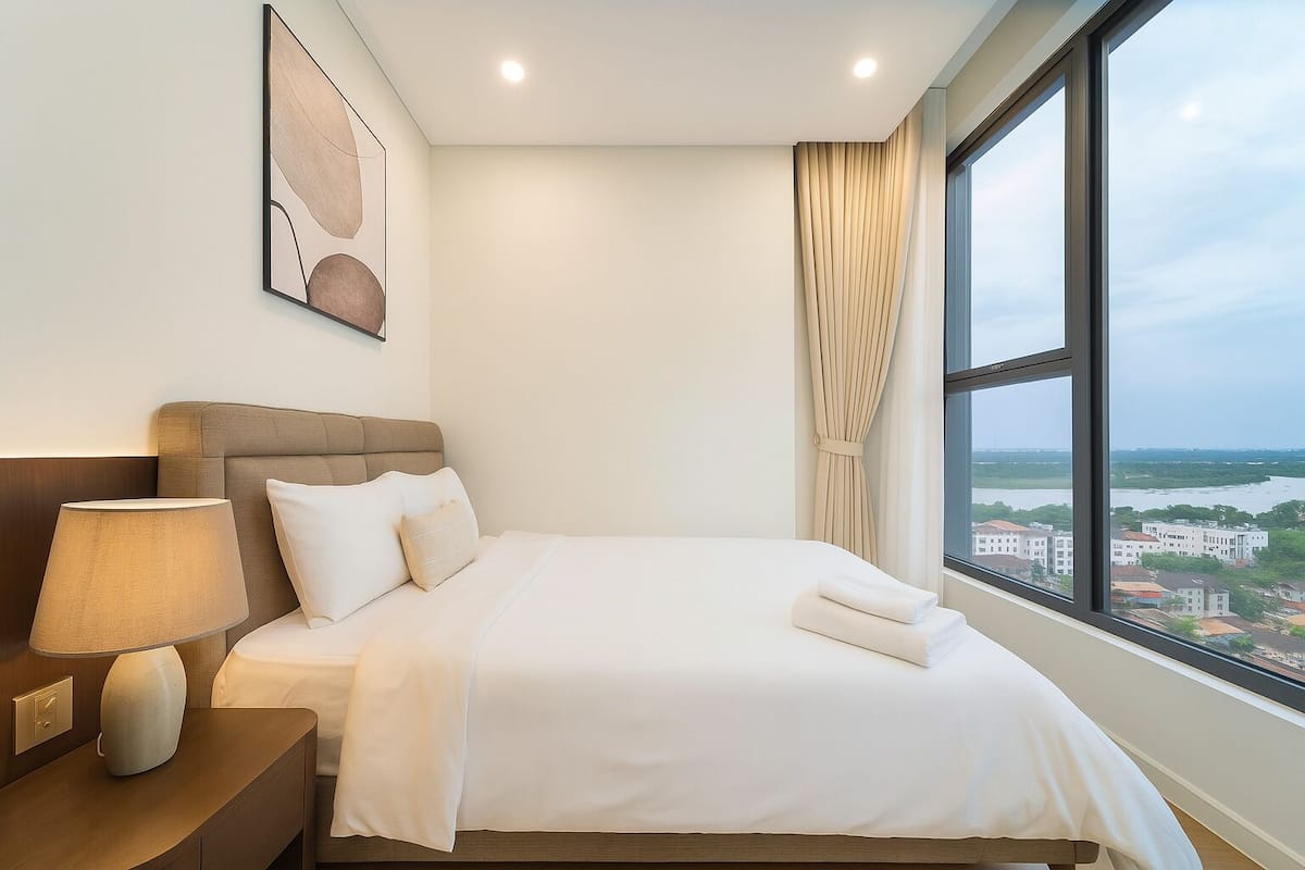 A hotel room with a bed, a nightstand, a lamp, a painting, and a window with a city view.
