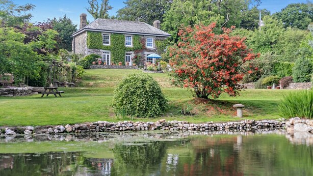 A stone house with ivy-covered walls, a pond, and a well-maintained garden with a variety of plants and a bench. - 3 Bed in Uphill (oc-n27747) (Linkinhorne)