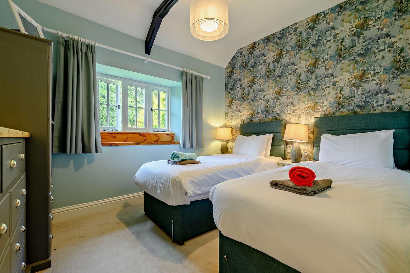 A hotel room with two beds, a dresser, a window with curtains, and a floral wallpaper.
