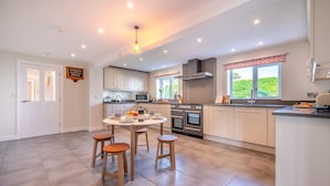 A modern kitchen with white cabinets, a central island, and a dining area with stools. - 6 Bed in Constantine Bay (oc-l27699) (Constantine Bay)