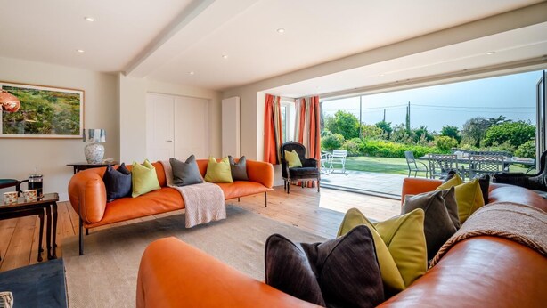 A spacious living room with a large orange sofa, a coffee table, and a view of a garden through large windows. - 6 Bed in Constantine Bay (oc-l27699) (Constantine Bay)