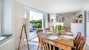 A dining area with a wooden table set for four, surrounded by chairs, with a view of a garden and a kitchen in the background. - 4 Bed in Mynytho (oc-b27888) (Mynytho)