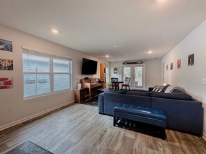 Ein Wohnzimmer mit einem dunkelblauen Sofa, einem Holztisch und einem an der Wand befestigten Flachbildfernseher. Es gibt eine Essecke mit Tisch und Stühlen sowie eine Küchenzeile mit Arbeitsplatte und Schränken.