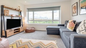 Ein Wohnzimmer mit großem Fenster, einem grauen Sofa, einem Flachbildfernseher, Holzregalen und einem gemusterten Teppich.