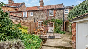 A stone house with a tiled roof, a wooden fence, and a garden with plants and a table. - 3 Bed in Wells-next-the-Sea (oc-c27939) (Wells-next-the-Sea)