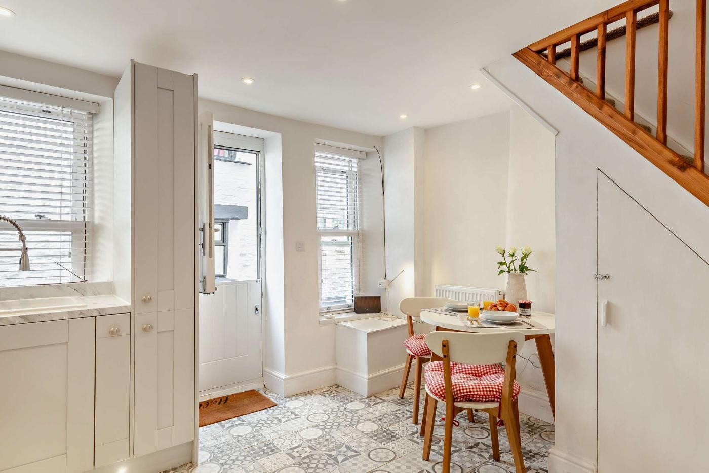 A compact kitchen with a dining area, white cabinetry, a wooden staircase, and patterned floor tiles.