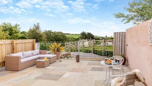Une terrasse avec un canapé, une table et des chaises, entourée d’une clôture en bois et offrant une vue sur la campagne.