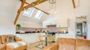 Un salon chaleureux avec un plafond haut et des fenêtres de toit, une cuisine avec des meubles blancs et un mur de fond en briques, et un coin repas avec une table et des chaises.