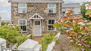 Une maison en pierre avec une porte centrale, de nombreuses fenêtres et un jardin où poussent différentes plantes et fleurs.