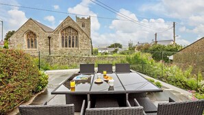 Un espace repas avec une table et des chaises, un bâtiment en pierre avec une tour en arrière-plan, et un jardin avec des fleurs et des arbustes.