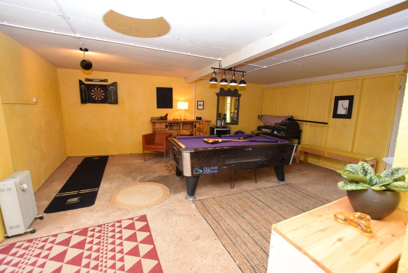 A room with a pool table, a dartboard, a chair, a small table with a plant, and a radiator.