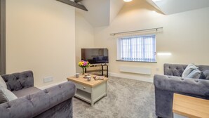 A modern living room with a flat-screen TV, two grey sofas, a wooden coffee table, and a window with blinds. - 2 Bed in Abersoch (oc-r26047) (Abersoch)