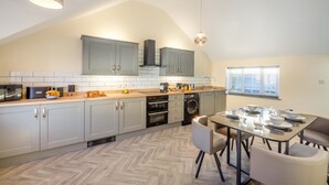 A modern kitchen with grey cabinets, a built-in oven, a washing machine, and a dining area with a table and chairs. - 2 Bed in Abersoch (oc-r26047) (Abersoch)