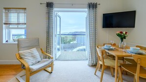 A modern living room with a sofa, dining table, and chairs. There is a large window with blinds, a flat-screen TV mounted on the wall, and a balcony with a view. - 2 Bed in Penally Near Tenby (oc-g28130) (Penally)