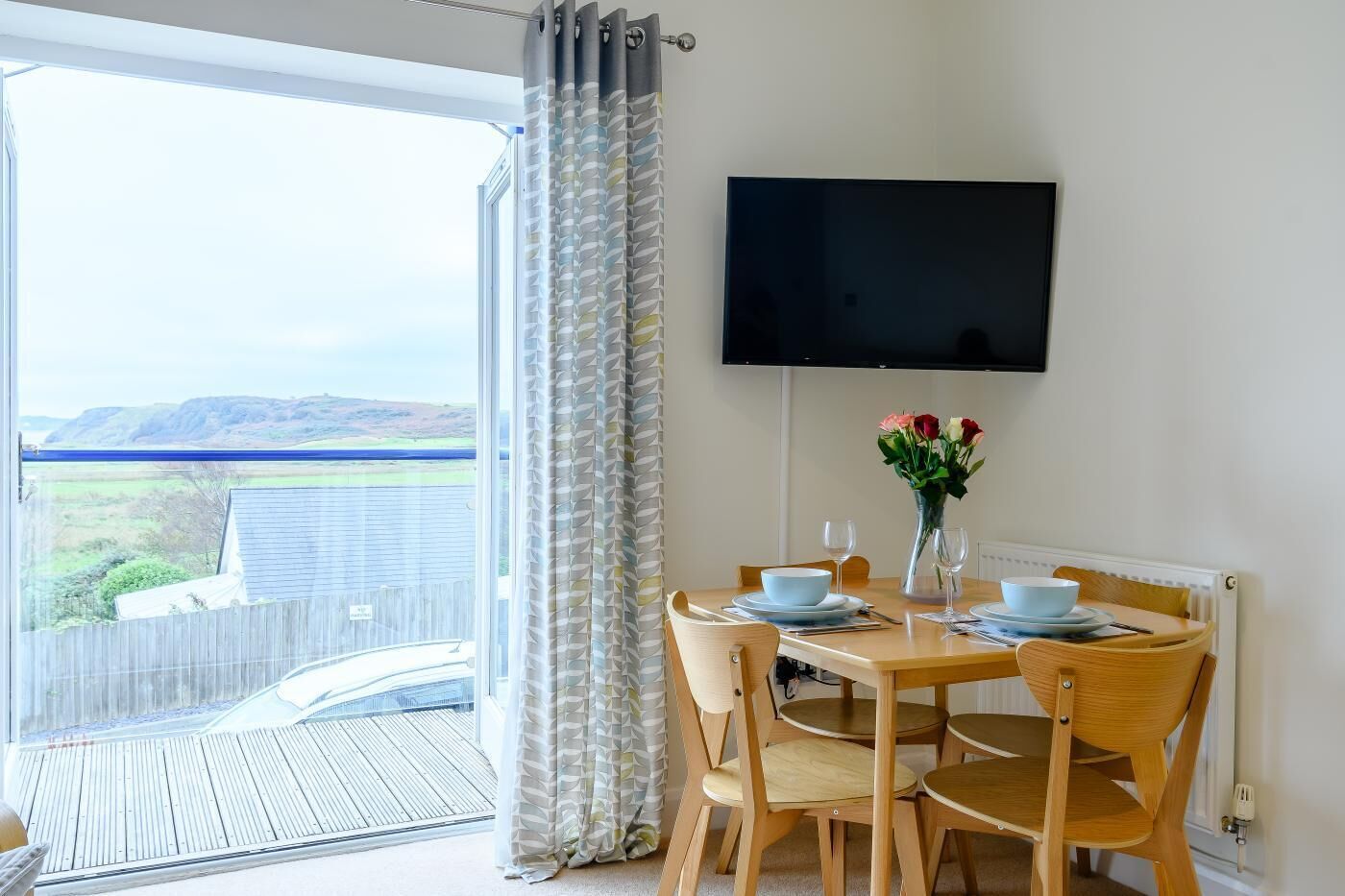 Un comedor con una mesa dispuesta para dos personas, un televisor de pantalla plana en la pared y vistas al exterior a través de un amplio ventanal.