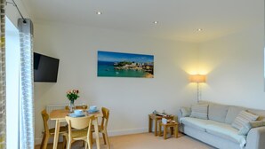 A living room with a dining area, a sofa, a TV mounted on the wall, and a painting of a coastal scene. - 2 Bed in Penally Near Tenby (oc-g28130) (Penally)