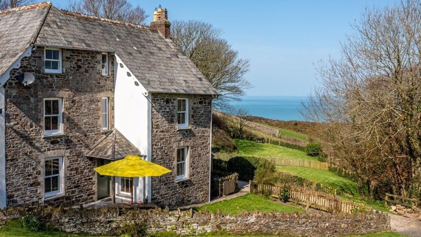 Ein Steinhaus mit weißer Tür und Fenstern, einem gelben Sonnenschirm und Blick auf das Meer.