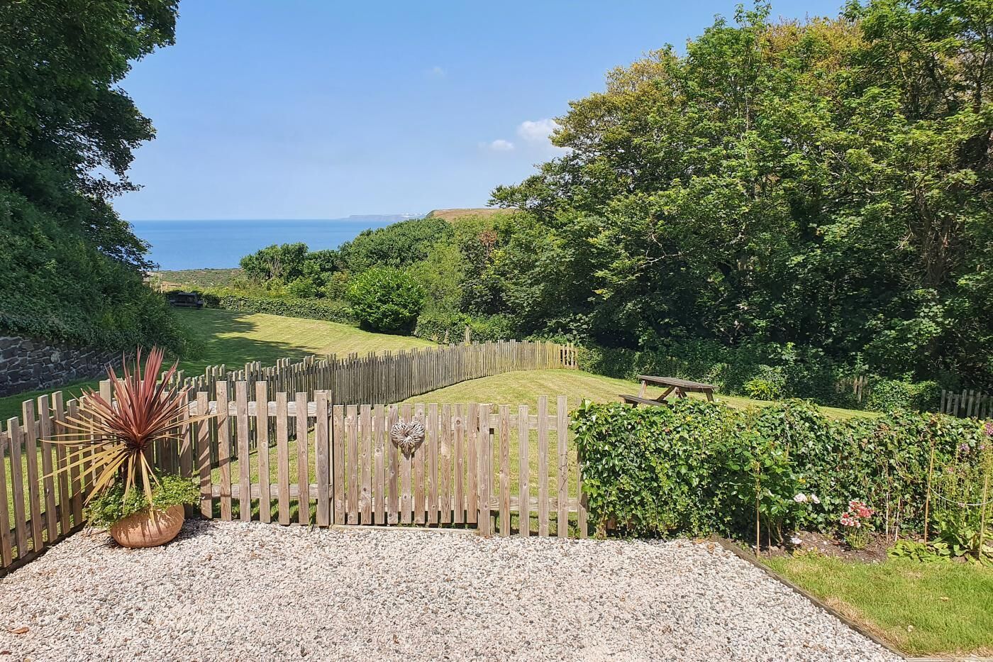 Une allée de gravier mène à un portail en bois orné d’un motif en forme de cœur, le tout étant entouré d’une végétation luxuriante et offrant une vue sur la mer.