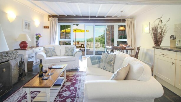 A cozy living room with a sofa, a coffee table, and a dining area with a table and chairs. There is a large window with a view of the outdoors. - 2 Bed in St. Gennys (oc-f28163) (Crackington Haven)
