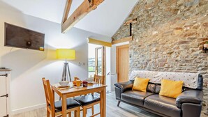 A cozy living room with a stone wall, a leather sofa with yellow cushions, a wooden dining table with chairs, and a flat-screen TV mounted on the wall. - 1 Bed in Morwenstow (oc-r28167) (Woodford)