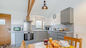A modern kitchen with a microwave, oven, and sink. There is a dining table with breakfast items and a clock on the wall. - 1 Bed in Morwenstow (oc-r28167) (Woodford)