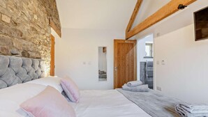 A bedroom with a stone wall, a wooden ceiling, a bed with pillows, a mirror, and a bathroom with a towel rack. - 1 Bed in Morwenstow (oc-r28167) (Woodford)