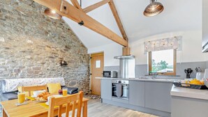 A modern kitchen with a stone wall, wooden beams, and a breakfast setup with a table and chairs. - 1 Bed in Morwenstow (oc-r28167) (Woodford)