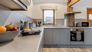 A modern kitchen with grey cabinetry, a built-in oven, a microwave, and a window with a view of the outside. - 1 Bed in Morwenstow (oc-j28166) (Woodford)