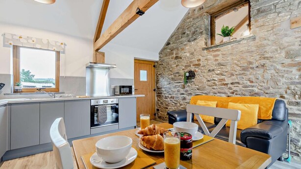 A cozy kitchen with a stone wall, wooden beams, and a dining area with a table set for breakfast. - 1 Bed in Morwenstow (oc-j28166) (Woodford)