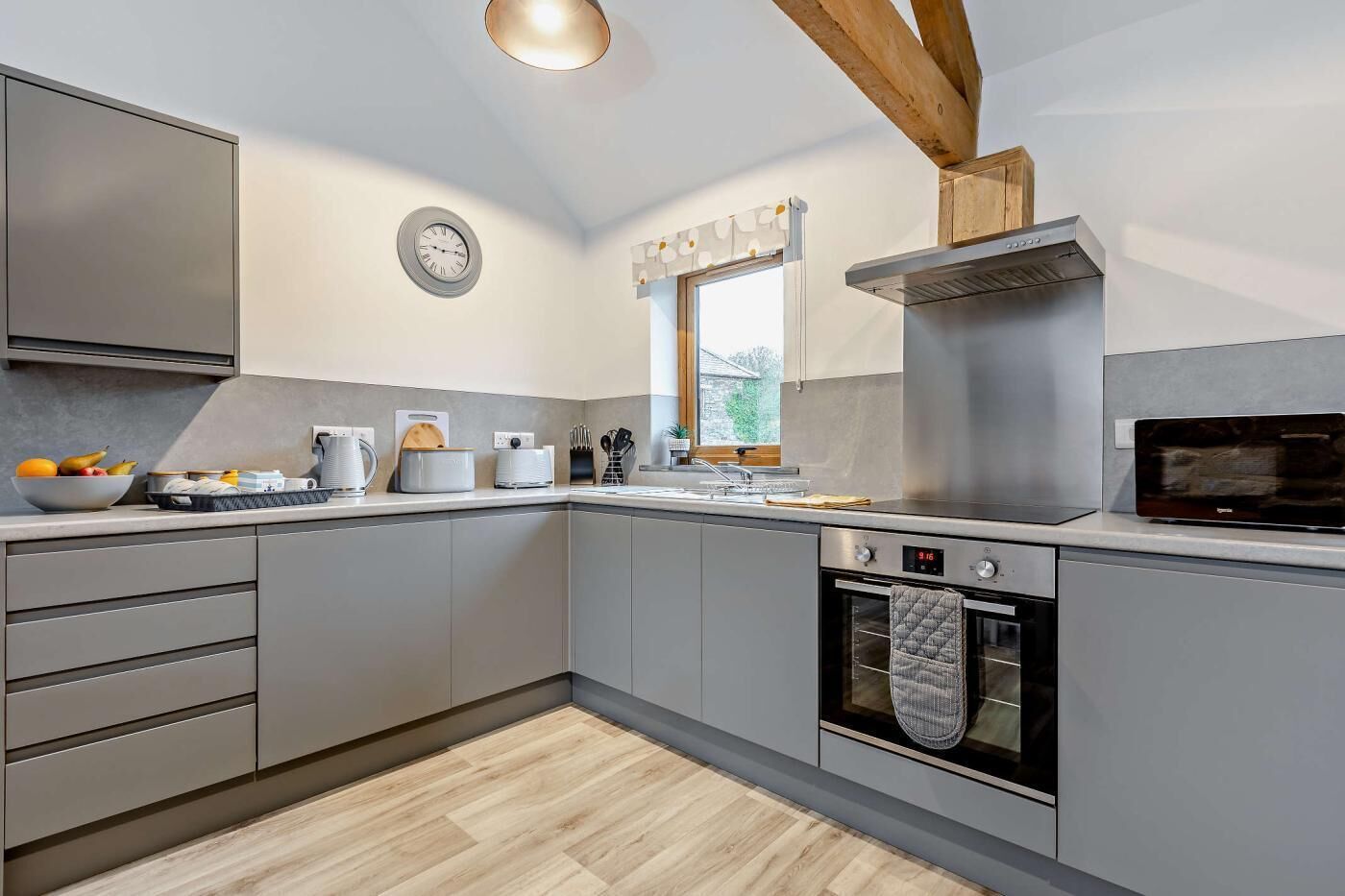 A modern kitchen with grey cabinets, a stainless steel range hood, a microwave, and a clock on the wall.