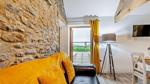 A room with a stone wall, a yellow sofa, a black leather bench, a tripod lamp, and a TV on the wall. - 1 Bed in Morwenstow (oc-j28166) (Woodford)