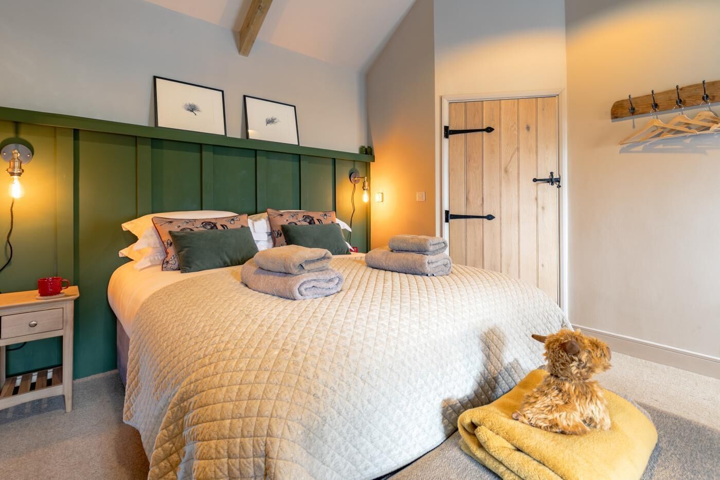 A bedroom with a bed, pillows, a bedside table, a wall-mounted shelf, a coat rack, and a dog on a blanket.