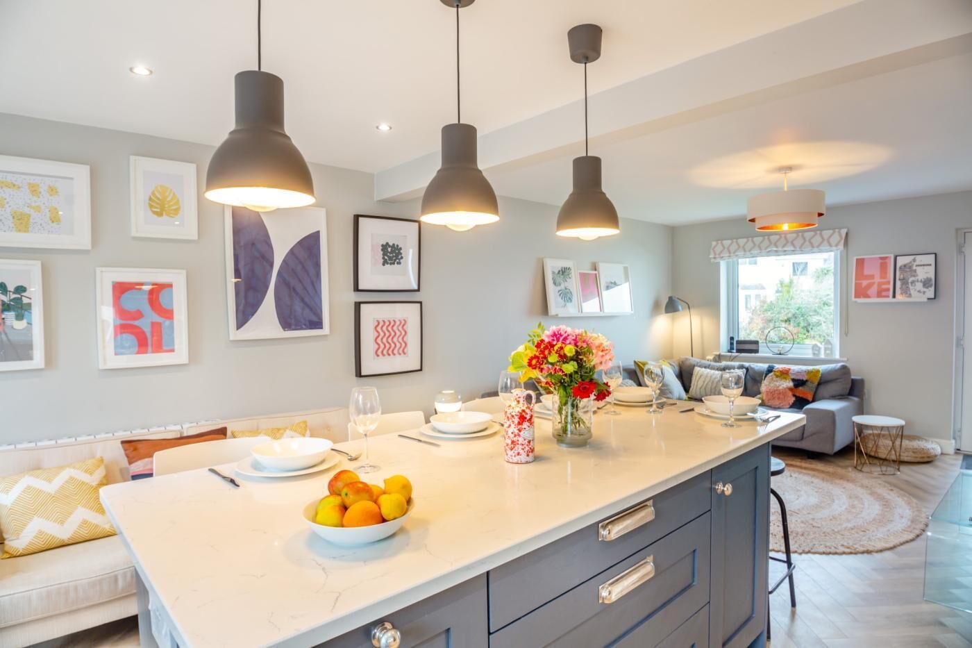 Een moderne keuken met hanglampen, een centraal eiland met fruitschaal, en een zithoek met een bank en tafel.
