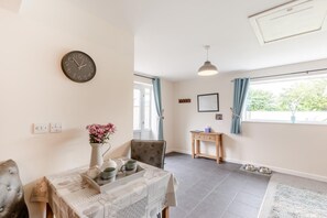 A room with a dining table, a clock, a small desk, a window with curtains, and a pet feeding station. - 1 Bed in Kilkhampton (oc-s28185) (Kilkhampton)