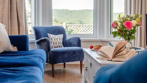 Une chambre à coucher avec deux lits bleus, un fauteuil bleu assorti, une commode avec un livre et une assiette de fraises, et une fenêtre donnant sur un paysage verdoyant.