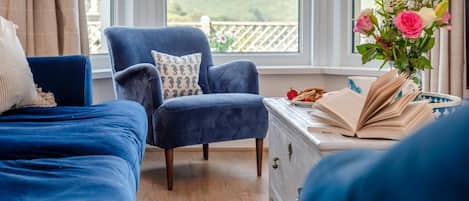 A bedroom with two blue beds, a matching blue armchair, a dresser with a book and a plate of strawberries, and a window with a view of greenery.
