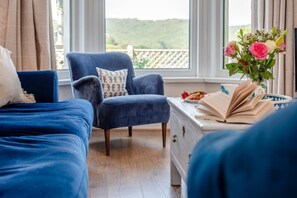 A bedroom with two blue beds, a matching blue armchair, a dresser with a book and a plate of strawberries, and a window with a view of greenery. - 2 Bed in Looe (oc-v28787) (Looe)