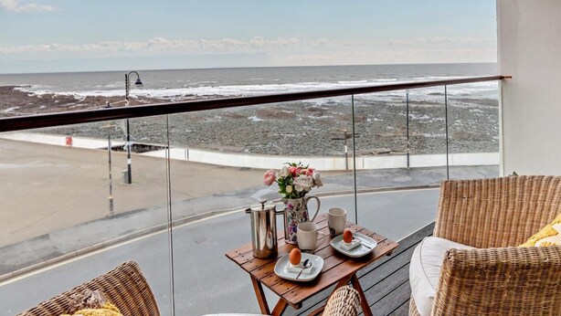 Un balcón con mobiliario de mimbre, una mesa con un jarrón de flores, una cafetera y una taza. Se ve el océano y una costa rocosa.