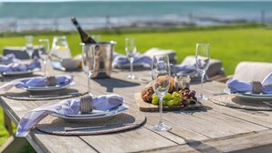 Una mesa de madera dispuesta para una comida, con platos, copas y una botella de champán, con vistas a una zona verde y al mar.